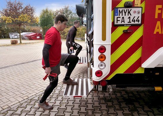 Zwei Personen in Sportkleidung mit Gewichten in den Händen haben jeweils ein Bein auf die seitelich herausgeklappten Trittbretter eines Feuerwehrfahrzeugs gestellt.