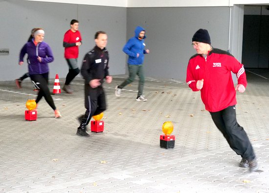 Mehrere Personen in Outdoor-Sportkleidung laufen Slalom um auf dem Boden aufgestellte Feuerwehr-Warnlampen.