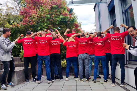 Eine Personengruppe mit gleichen T-Shirts mit der Rückenaufschrift "Feuerwehrsport-Coaches". Sie stehen mit dem Rücken zum Betrachter und zeigen zusammen mit zwei Übungsleitern, die jeweils an einer Seite stehen, auf ihre T-Shirts.