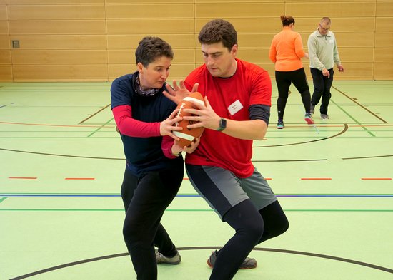 Zwei Personen in einer Sporthalle bei einer Übung. Sie stehen Rücken an Rücken und geben sich über eine Seite einen Rugby-Ball an. Im Hinergrund weitere sporttreibende Personen.