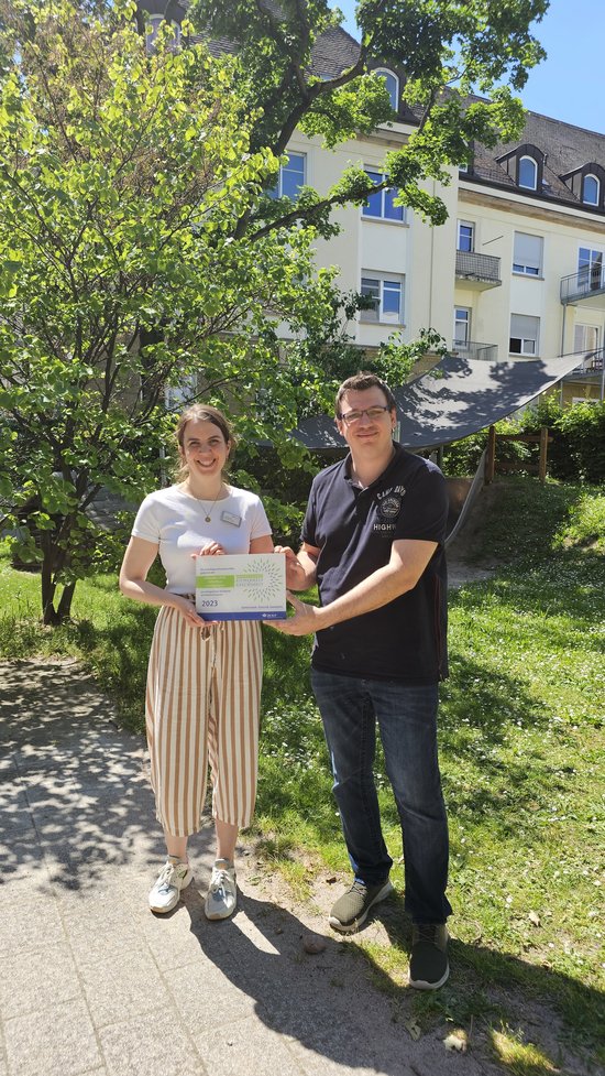 Zwei Personen im Außenbereich einer Kita. Beide halten gemeinsam die Präventionspreisplakette in den Händen.