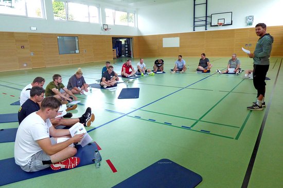 Eine Sportgruppe sitzt auf Matten in einer Sporthalle in einem Halbkreis um einen Trainer herum. Alle haben ein Blatt Papier in der Hand.
